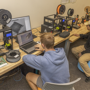 Two students sit facing laptops and 3D printers. A red banner over part of the image includes the words 
